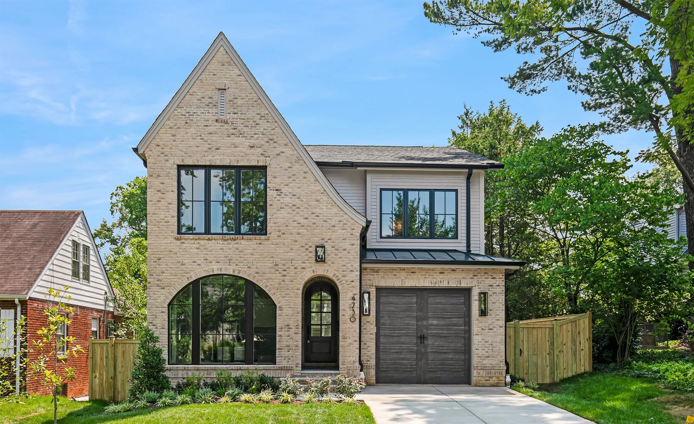 Charming two-story brick new construction home in Arlington, VA with steep gable roof, arched front windows and doorway, black-framed windows, and a single dark wood garage door, surrounded by green lawn and mature trees.