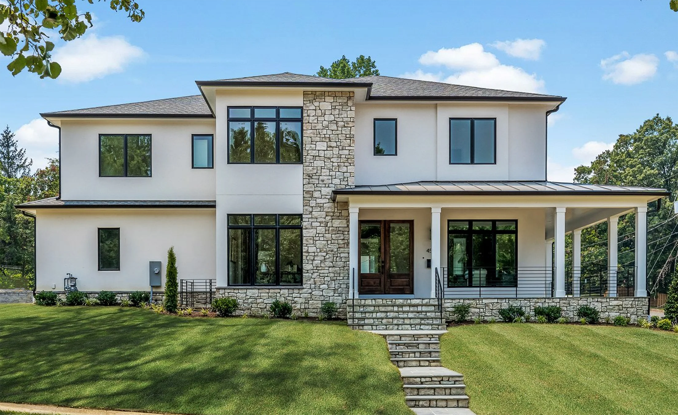 Contemporary Arlington new construction custom luxury two-story home with white stucco exterior, central stone accent wall, black-framed windows, and a covered front porch with modern railing and double wood front doors, surrounded by manicured lawn and trees.