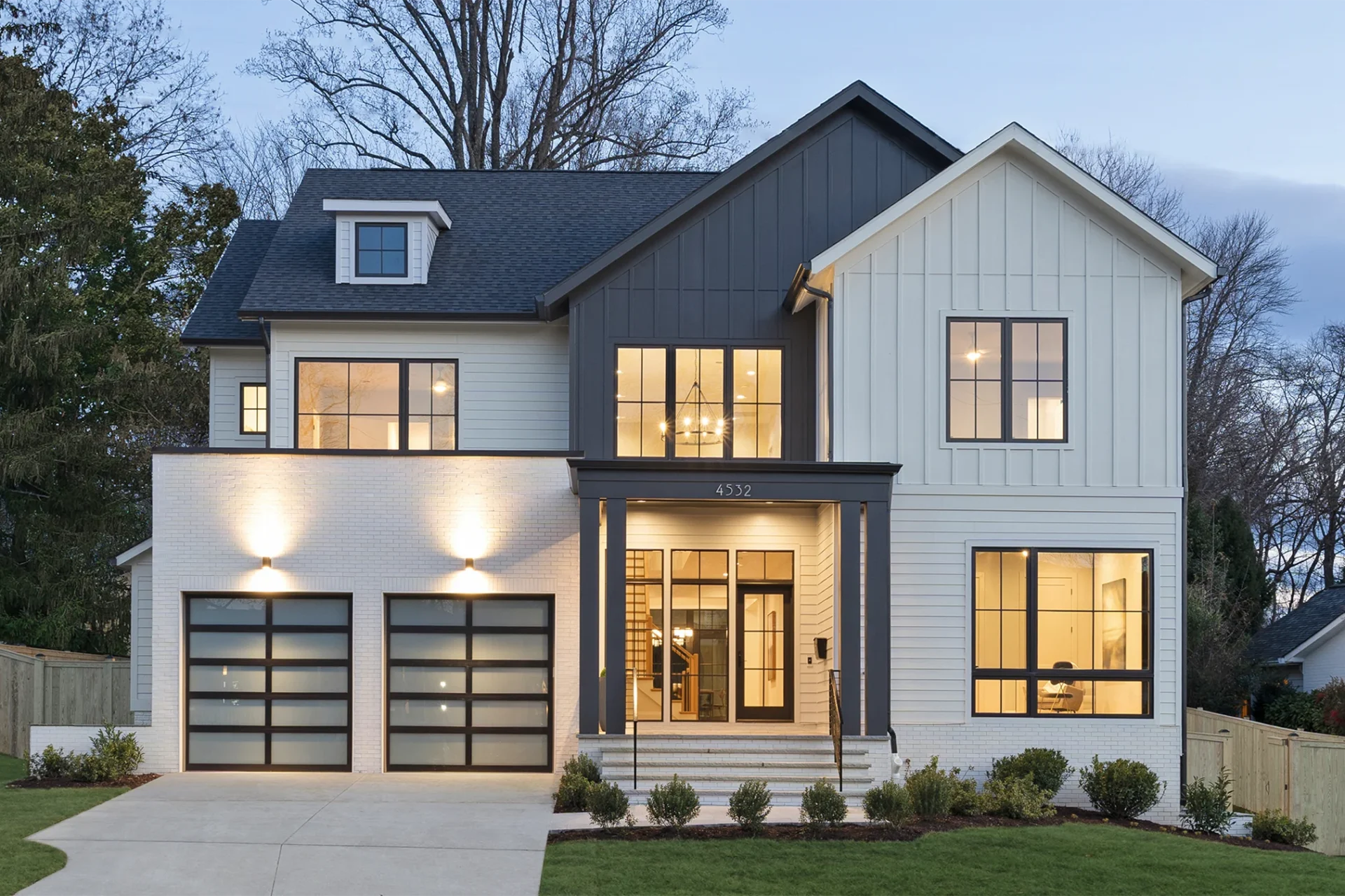 Modern two-story farmhouse-style new custom home with white board-and-batten siding, dark gray gable accents, large black-framed windows, and double glass garage doors, illuminated by warm exterior lighting at dusk.