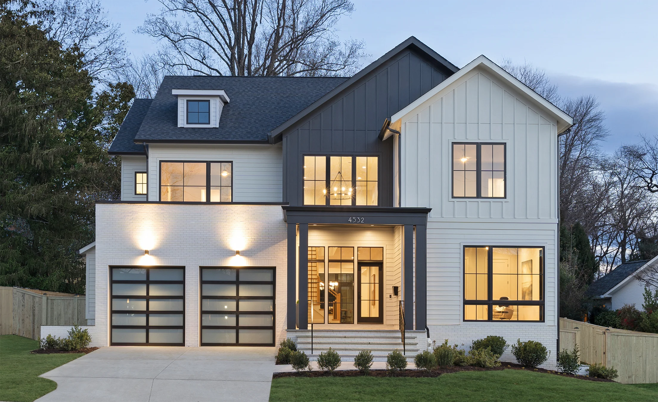 Modern two-story farmhouse-style new custom home with white board-and-batten siding, dark gray gable accents, large black-framed windows, and double glass garage doors, illuminated by warm exterior lighting at dusk.