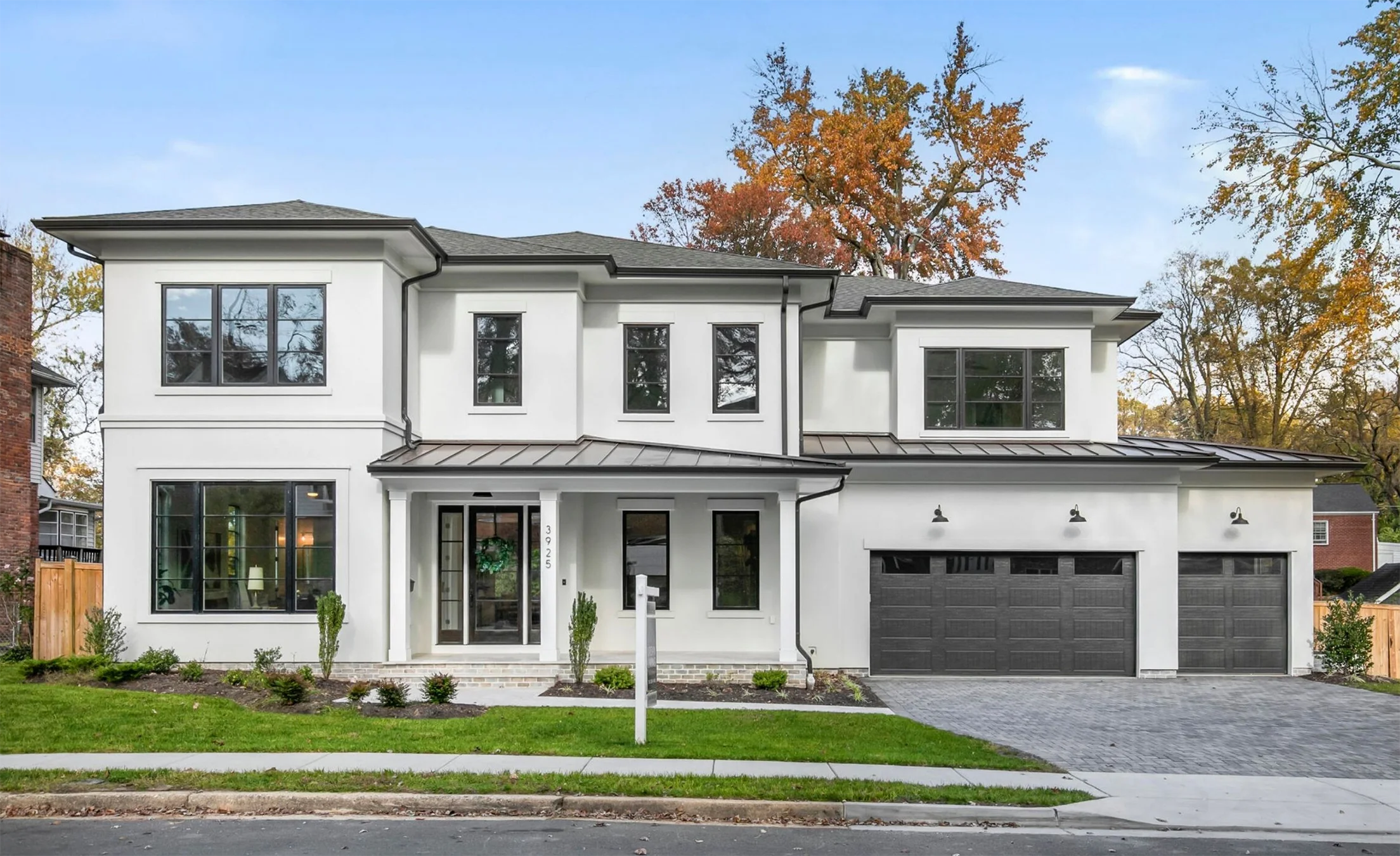 Modern luxury new construction two-story home with white stucco exterior, black-framed windows, dark metal roof accents, and a three-car garage with black doors, featuring a covered front entry and landscaped front yard with green lawn and shrubs.